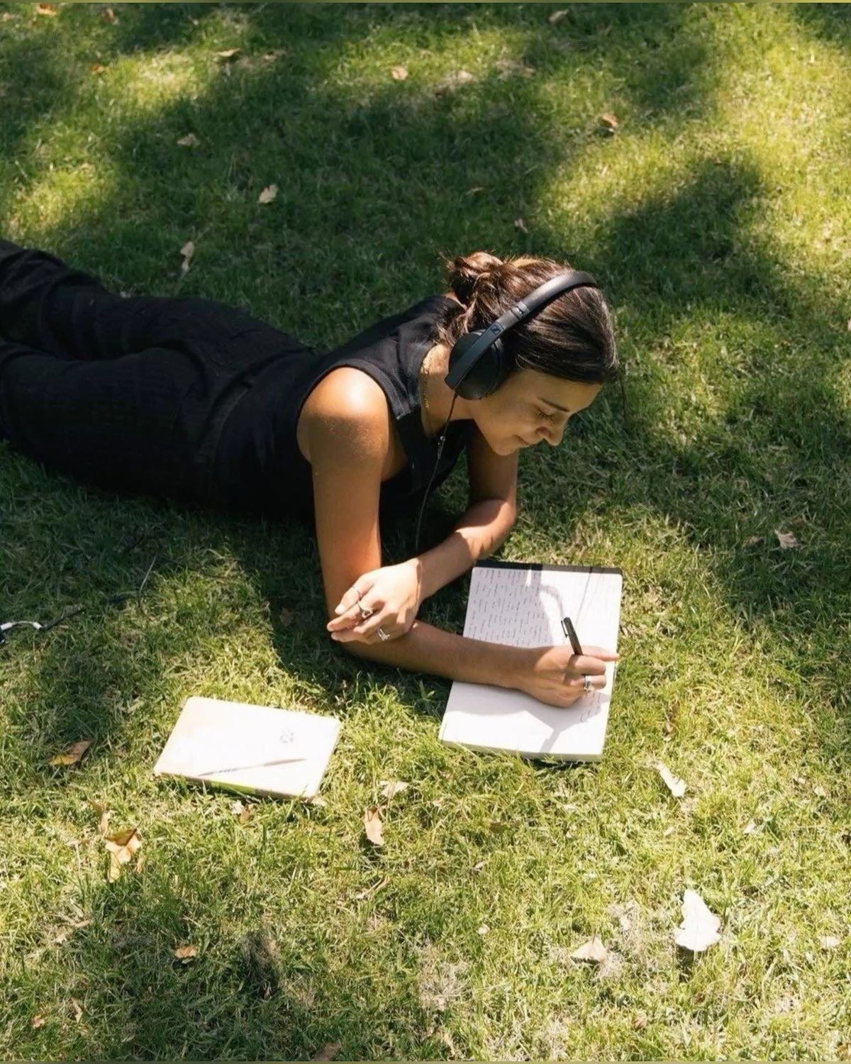 Una persona joven tumbada boca abajo sobre el césped de un parque, disfrutando de un momento de introspección y calma. Viste ropa oscura y lleva unos auriculares grandes puestos mientras escribe con concentración en un cuaderno abierto. A su lado, sobre la hierba iluminada por el sol, descansa otro libro pequeño. La escena transmite una atmósfera de serenidad, estudio o creatividad al aire libre.